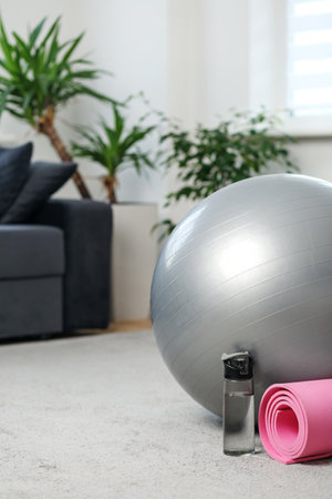 Minimalist home gym scene with a silver stability ball, pink yoga mat, and water bottle on a carpeted floor near sofa.の写真素材
