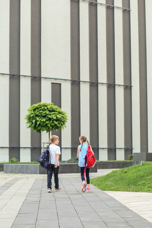 Two cheerful children with backpacks go along a paved path outside a modern school building, enjoying their break or arrival.の写真素材