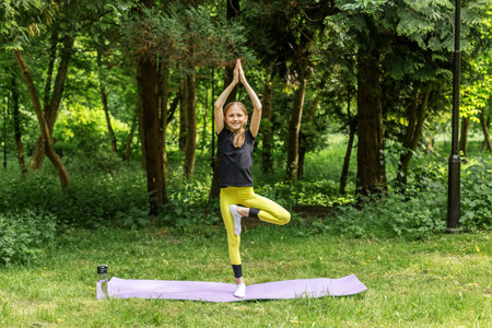 Young girl practices yoga tree pose on mat in forest setting, showing balance, mindfulness, and enjoyment of nature.の写真素材