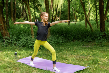 Tween girl in athletic wear performs warrior pose on yoga mat in a green forest setting, focusing on balance and mindfulness.の写真素材