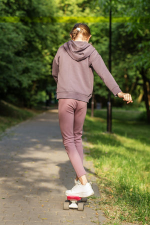 Preteen girl practices balance while riding a skateboard along a quiet, tree-lined park pathway on a sunny day.の写真素材