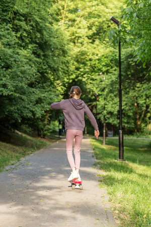 Tween girl practices balance while riding a skateboard along a quiet, tree-lined park pathway on a...