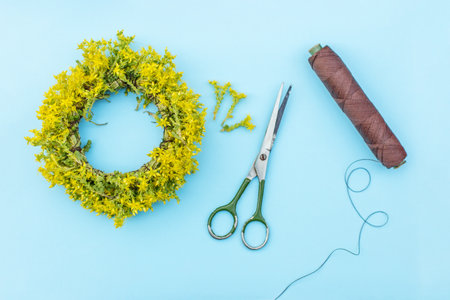 Flat lay of yellow flower wreath, green-handled scissors, and brown thread on a light blue surface, ready for crafting.の写真素材