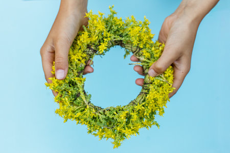 Close-up of two hands presenting a small circular wreath made of fresh yellow flowers, with a pastel blue background.の写真素材