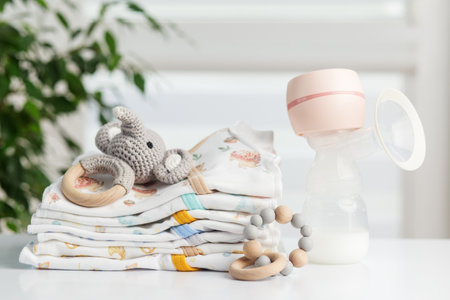 Electric breast pump with milk bottle placed beside folded baby clothes, a crochet elephant toy, and...