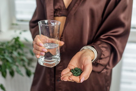 Close-up of woman's hands showing a handful of green tablets and a glass of water, representing daily supplements and wellness routine.の写真素材