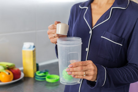 Close-up of a woman in pajamas holding a scoop of protein powder above a shaker cup in a modern...