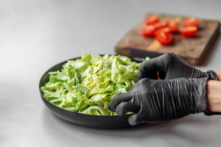 Person wearing black gloves assembling a Caesar salad with fresh romaine lettuce, with sliced tomatoes blurred in the background.の写真素材