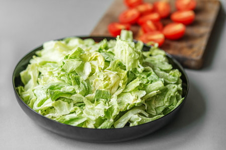 Close-up of a fresh salad with romaine lettuce in a black bowl, with sliced cherry tomatoes in the background.の写真素材