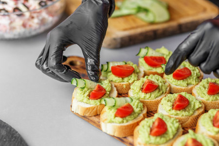 Gloved hands placing fresh cucumber and tomato on avocado-topped baguette slices, creating colorful and healthy bite-sized appetizers.の写真素材