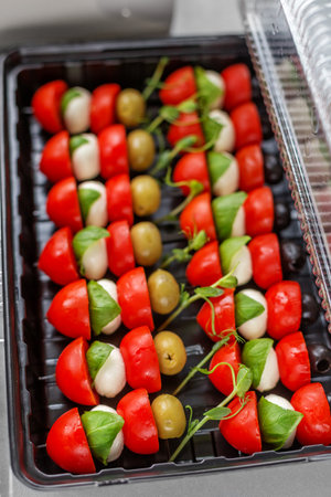 Fresh caprese-style skewers with mozzarella balls, cherry tomatoes, green olives, and basil leaves in a black tray for events.の写真素材