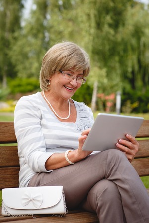 Attractive mature woman sitting on a bench holding and using a digital tablet in the park at summer dayの写真素材