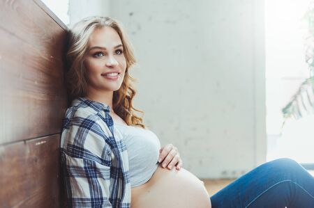 Happy pregnant woman sitting on the floor and touching her belly at homeの写真素材
