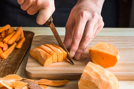 Young woman cutting with knife sweet potato into wedges, peels on wood table, sliced carrots, kitchen interior, close upの写真素材