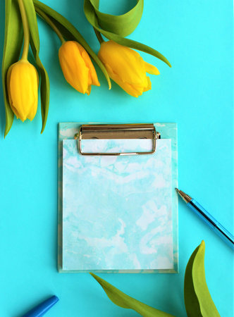Blank clipboard with yellow tulips on blue background, flat lay
