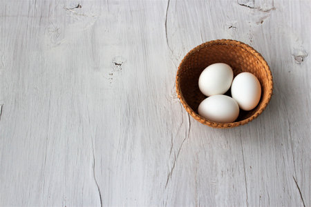 White eggs in a wicker basket on a white wooden background.