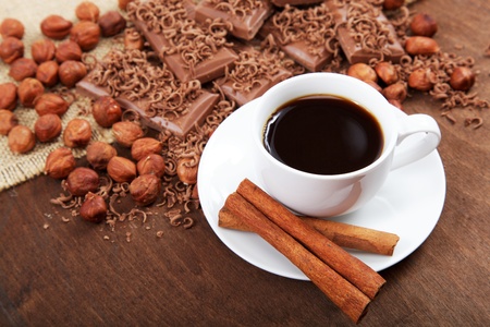A cup of coffee with cinnamon sticks in a saucer, chocolate and hazelnuts on a wooden table.の写真素材