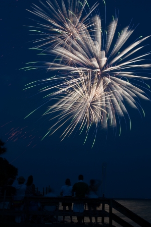Photograph of various firework bursts from a 4th of July Celebration on Saint Simons Island Georgia   Fireworks are shot off over water and reflection backlights people in foreground の写真素材
