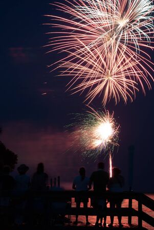 Photograph of various firework bursts from a 4th of July Celebration on Saint Simons Island Georgia   Fireworks are shot off over water and reflection backlights people in foreground の写真素材