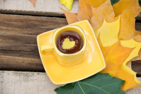 Yellow leaf is at the yellow cup of tea on the saucer with autumn leaves on a wooden table.の写真素材
