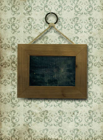 empty wooden frame fixed on a old and dirty wall, the inner part of the frame is a slate, the wallpaper is stained and dirty with arabesques on it, the frame is fixed by a nail ans a ropeの写真素材