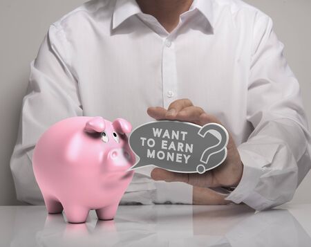 Image of a man hand holding speech balloon with the text want to earn money and pink piggy bank, white shirt. Concept for money earning.の写真素材