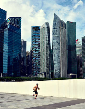 A muscular man, shirtless, jogging at the foot of buildings in Singaporeのeditorial素材