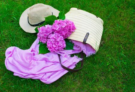 Pink hydrangea flowers in summer bag and sun hat on lush green grass.