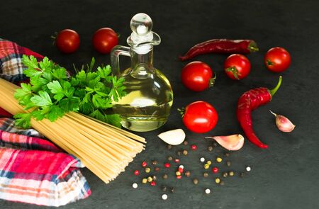 Ingredients for cooking spaghetti - raw spaghetti, cherry tomatoes, chili peppers, garlic, herbs, spices and olive oil on a black background. Culinary background. Selective focus.の写真素材