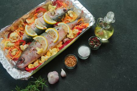 Baked fish with vegetables and lemon and spices in a baking dish. Top view on a black background....