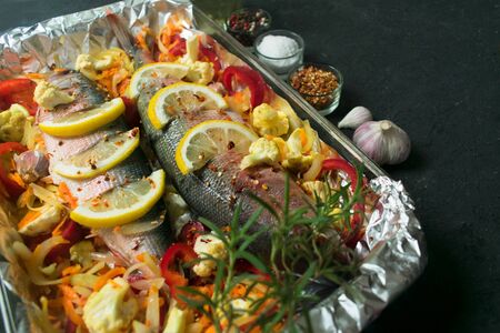 Baked fish with vegetables and lemon and spices in a baking dish. Top view on a black background. Close-up.の写真素材