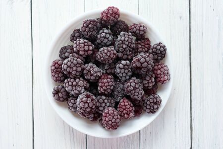 Frozen blackberries in a plate on a wooden white background. Top view.