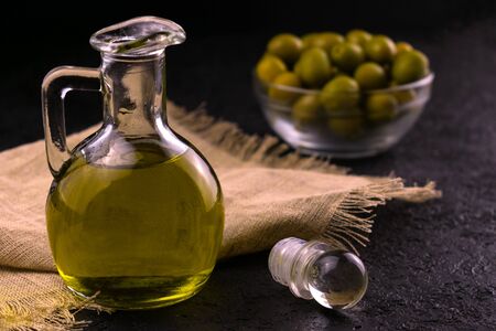 Olive oil in a jug on a black background and olives in a glass plate in the background. The concept of olive oil.の写真素材