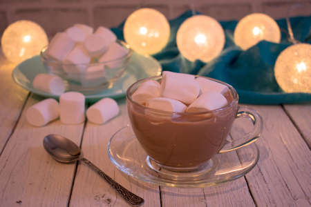 A cup of cocoa with marshmallows on a white wooden table against the background of Christmas lights.