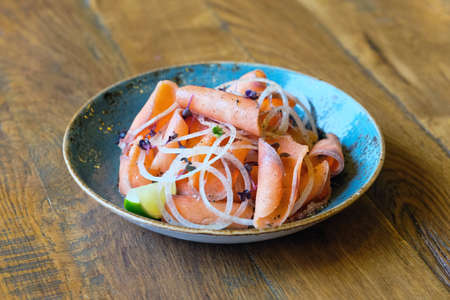 Red fish stroganina in a blue ceramic plate with onions and black pepper on a wooden table.の写真素材