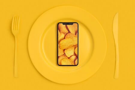 Order food online, food delivery. Call and bring food home. Smartphone with chips on the screen, lying on a plate, knife and fork. Yellow. The view from the top.の写真素材
