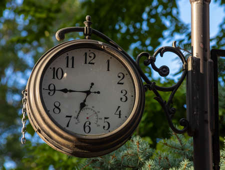 Old fashioned vintage street clock against greenl trees and blue sky.の写真素材
