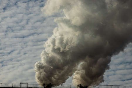 White and gray smoke and steam from a high concrete chimney against the bright blue sky.