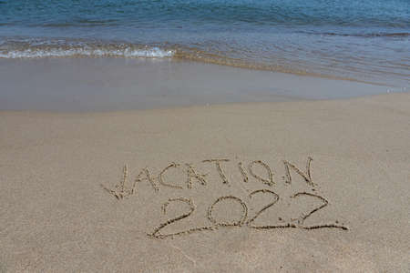 The inscription vacation 2022 on the sand by the water and the rising wave, seashore beach vacation...