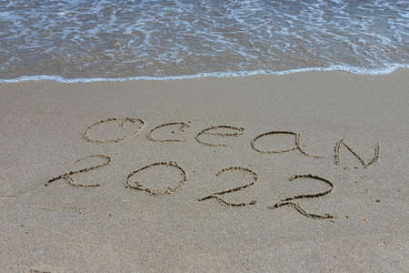 The inscription ocean 2022 on the sand by the water and the rising wave, seashore beach vacation by the sea.の写真素材
