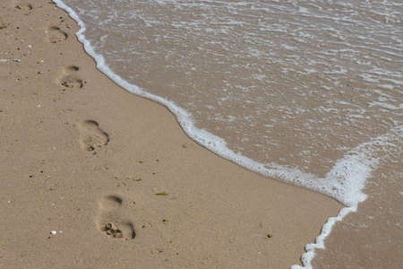 Footprints in the sand beach. Footprints in the sand against a sea wave. Footprints on a sunny day with golden sand, beach, wave and footsteps.の写真素材
