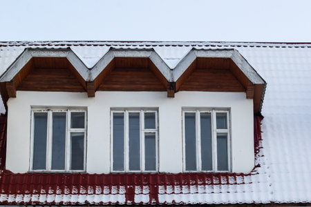Snow on the roof of a red, brown metal roof of a European house with a window.の写真素材