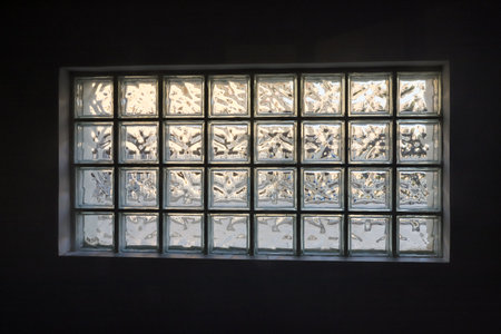 Soft light illuminates a textured glass block window, casting intricate patterns in the dim interior of a room as dawn breaks outside.の写真素材