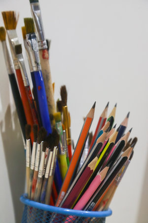 A variety of art supplies are arranged in a metal cup, showcasing vibrant colored pencils and paintbrushes against a plain white wall.の写真素材