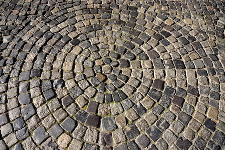 A circular design made of cobblestones forms an intricate pattern on the ground, set in a vibrant public square bordered by lush greenery and historical buildings.の写真素材