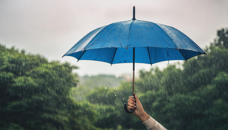 A hand emerges from the rain, gripping a blue umbrella while heavy raindrops cascade in a vibrant natural landscape during a stormy day.の素材