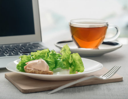 A light lunch featuring a plate of fresh salad and a slice of bread with a spread sits on a workspace next to a laptop and a cup of tea. The setting is calming and conducive to work.の素材