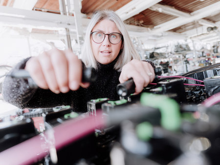 A skilled female mechanic is focused on replacing a car battery in a bustling workshop. She works efficiently with tools showing her expertise in auto repair.の素材