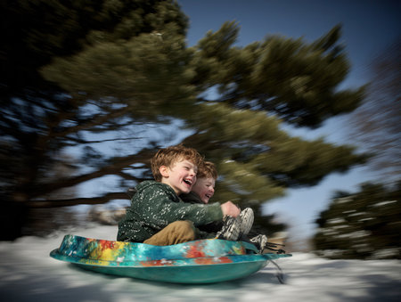 Two children laugh joyfully as they are quickly pulled on a colorful sled through the snowy landscape on a sunny winter day.の素材
