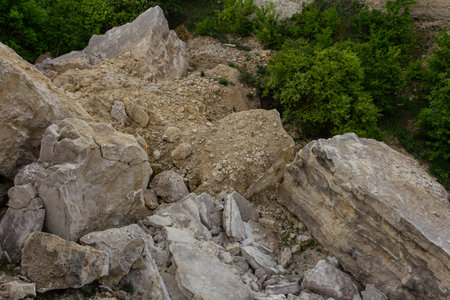 Large boulders and loose rocks create a dramatic landscape contrasting with the vibrant greenery of shrubs and trees nearby on a sunny day.の写真素材
